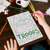 Woman sitting at brown table, coloring a Support Our Troops coloring page.