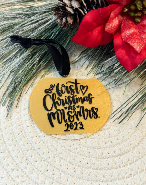 Gold painted acrylic ornament with hand lettered words saying “our first christmas as Mr. And Mrs. 2023” written on in. Also has the year 2023 as well as a beautiful black velvet ribbon.
