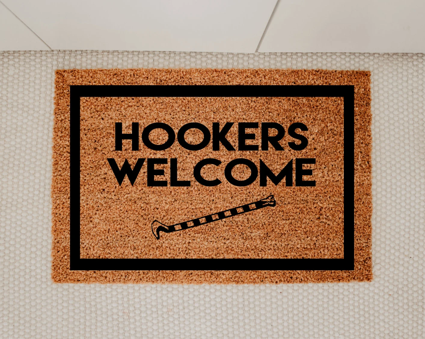 Entire design black. Centered block letters read hookers welcome. One word above the other. Below text and centered is image of tail hook from navy aircraft. Border 1.5 inch thick all the way around edge of mat. Mat is brown coconut husk.
